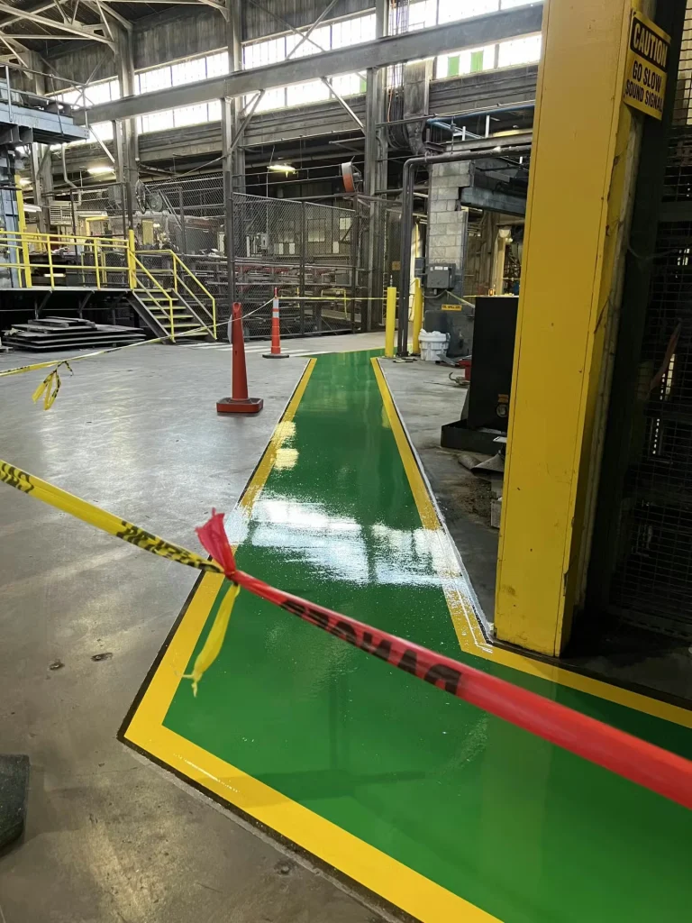 Narrow corridor in an industrial area with yellow guardrails flanking a shiny green walkway bordered in yellow; equipment and workstations line the sides.