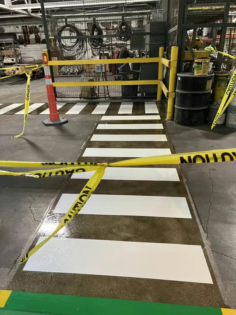 Close view of a white zebra crosswalk on dark concrete, glossy from fresh coating; the crosswalk leads to a green lane bordered in yellow near yellow railings.