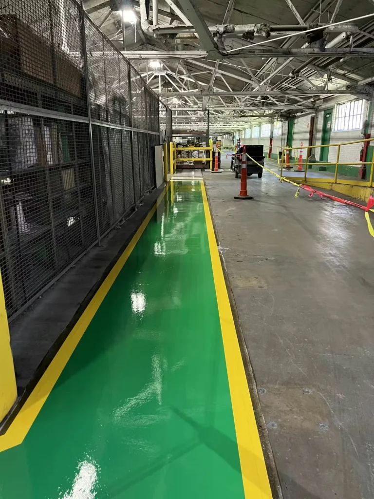 Wide warehouse view showing a glossy green pedestrian lane bordered in yellow extending forward; caution tape stretches across the lane and orange cones stand nearby.