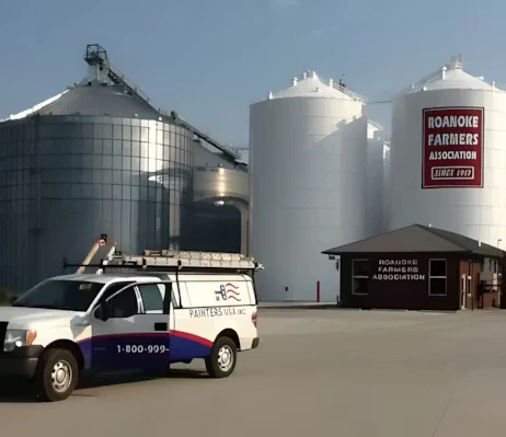 A white-and-blue painting company van is parked in front of large metal grain silos at an industrial site; a tall sign on one silo reads “Roanoke Farmers Association,” and a small office building sits to the right under a clear sky.