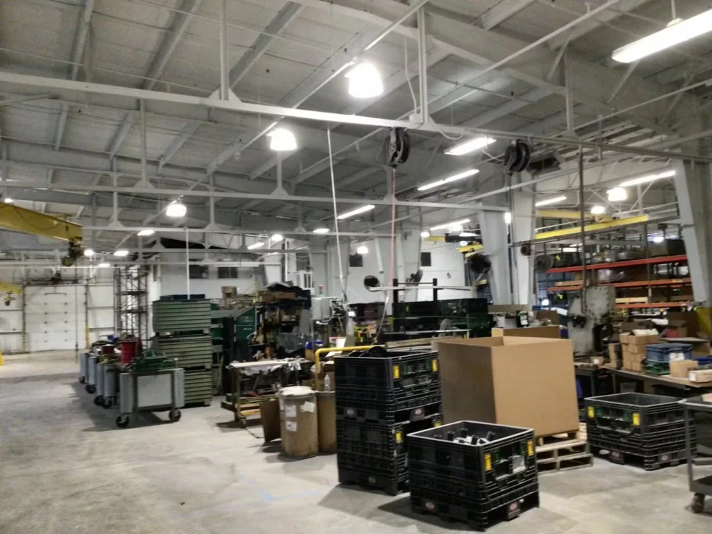 Wide view of an industrial workshop with a newly painted light-colored ceiling, bright overhead lights, and bins, pallets, and equipment on the floor.