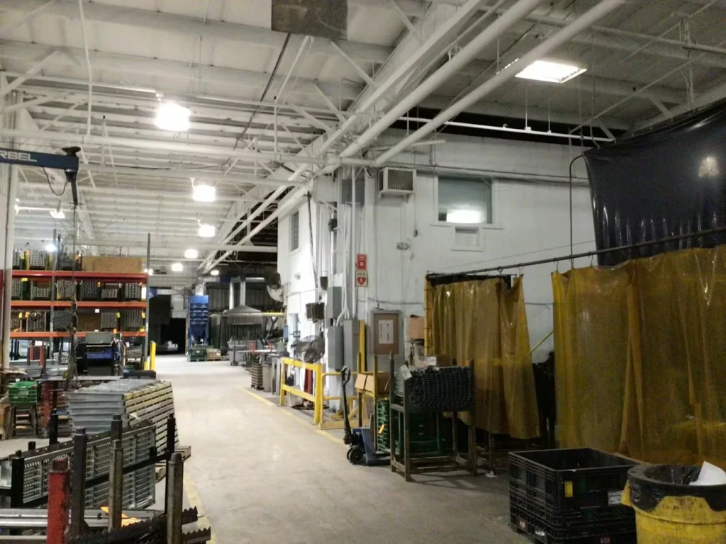 Factory interior with tall storage racks and a welding area screened by dark curtains; new ceiling paint and suspended lights hang above stacked boxes and materials.