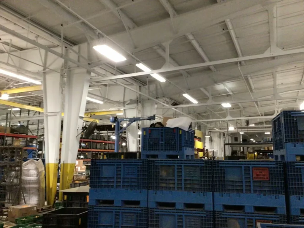 Upward view of a high, freshly painted ceiling with exposed beams, hanging lights, and ductwork above an industrial production area.