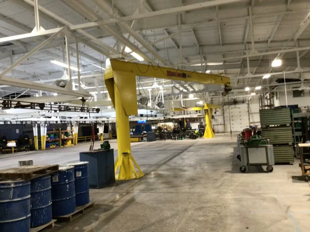 Two yellow overhead jib cranes stand over worktables and bins in a manufacturing area; bright lights and freshly painted ceiling beams fill the background.