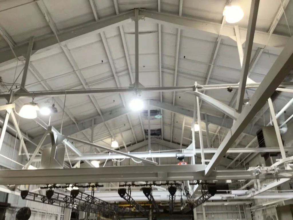 Warehouse section with stacked blue plastic pallets and tall white columns; overhead fluorescent fixtures and newly painted ceiling framing are visible.