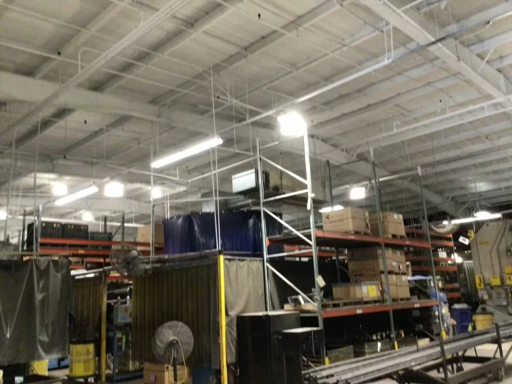 Long aisle inside an industrial facility with racks of parts on the left and yellow safety curtains on the right; bright lights and a freshly painted ceiling span overhead.