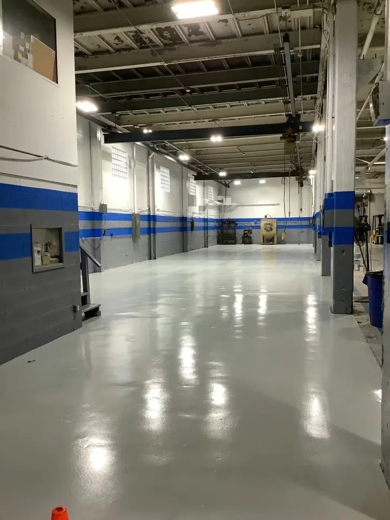 Empty industrial bay with a newly coated glossy light-gray floor reflecting overhead lights; white walls with blue and gray horizontal stripes and an overhead crane run down the space.