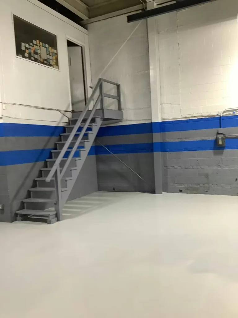 Factory interior corner with a small office door reached by gray metal stairs; walls have blue and gray stripes and the floor is freshly coated in smooth light-gray epoxy.