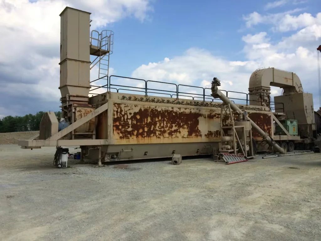 Large industrial processing machine outdoors on a gravel lot, with heavy rust staining along its side panels, ladders and guardrails on top, and ducting and piping attached; cloudy blue sky overhead.