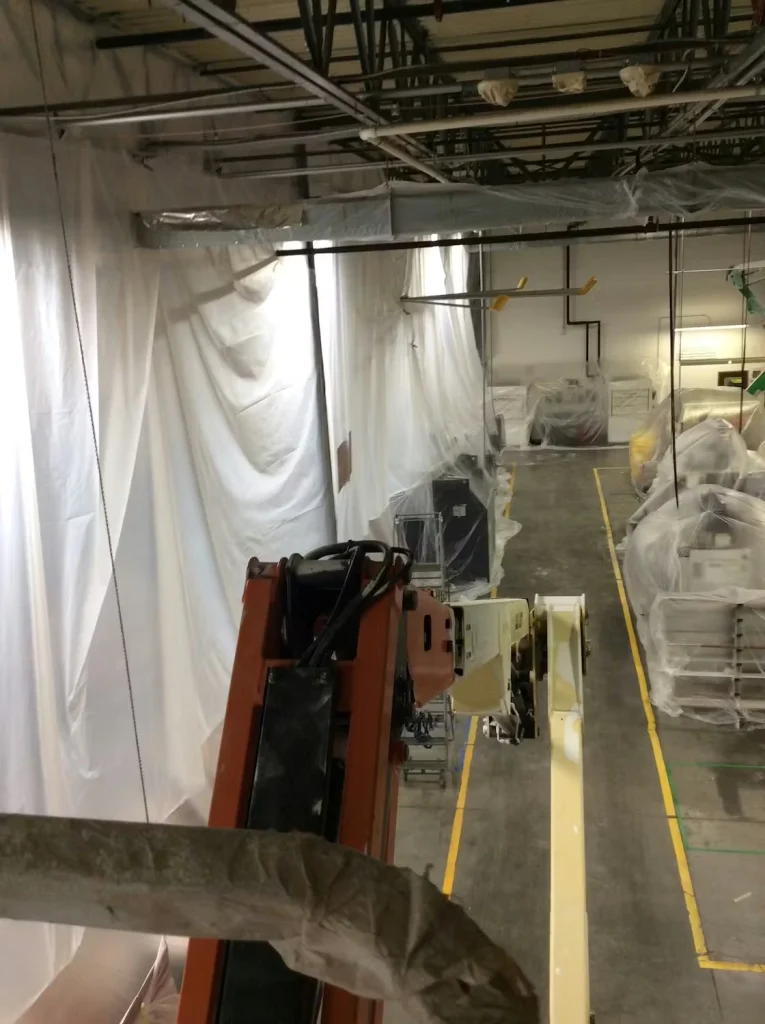 View from an elevated lift inside an industrial facility, with plastic sheeting hung along the walls and covering equipment below; exposed ceiling pipes and lighting run overhead, and yellow floor lines mark aisles.