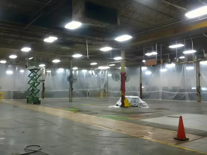 Large industrial shop floor enclosed with hanging plastic sheeting, with bright overhead lights, a green scissor lift, and a few cones marking areas; equipment is covered with tarps as work is underway.