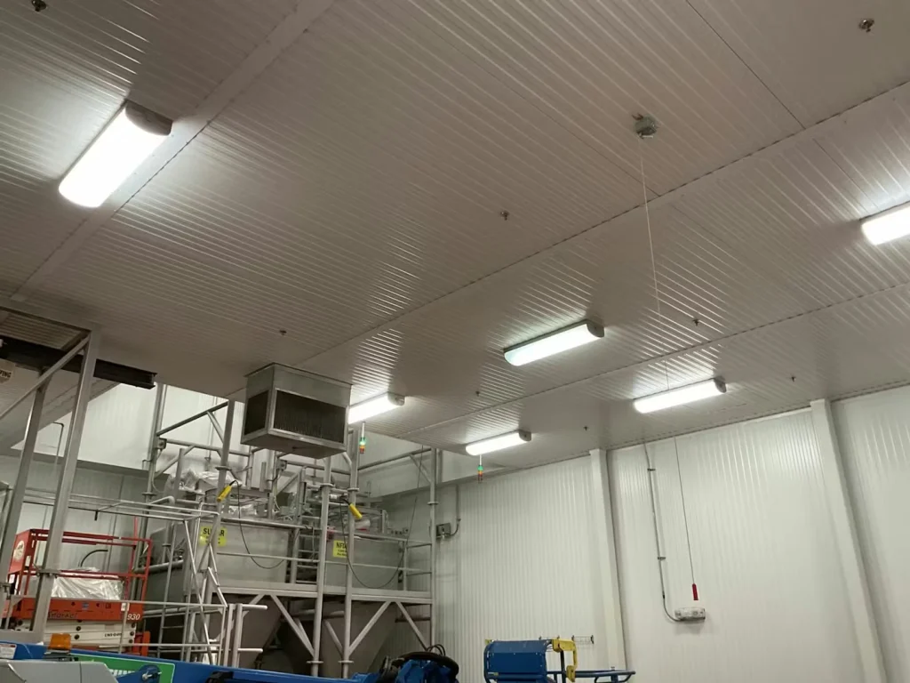 Interior of a clean industrial room with a white ribbed ceiling and bright fluorescent fixtures; stainless processing equipment and a scissor lift are visible below.