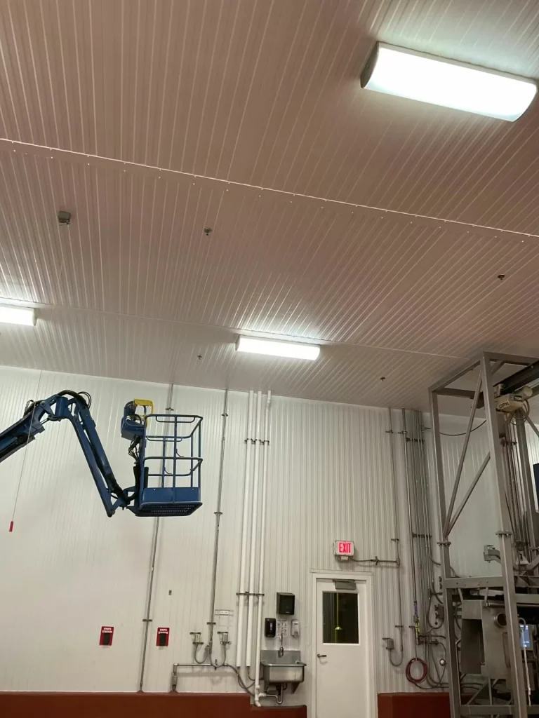 High-ceiling industrial space with white wall panels and a ribbed white ceiling lit by fluorescent lights; a blue boom lift basket is raised near the wall above an exit door and sink.