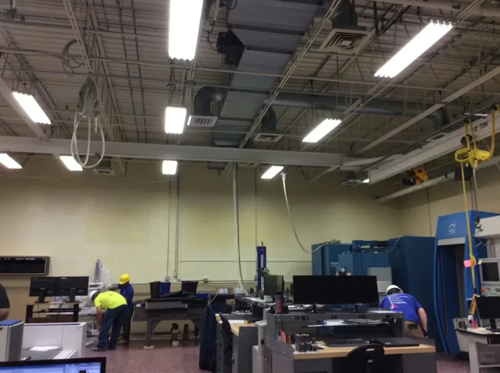 Industrial workspace with an exposed ceiling, ductwork, and bright fluorescent lights; two workers in hard hats and work shirts stand near machinery and computer stations, with large blue equipment along the right wall.