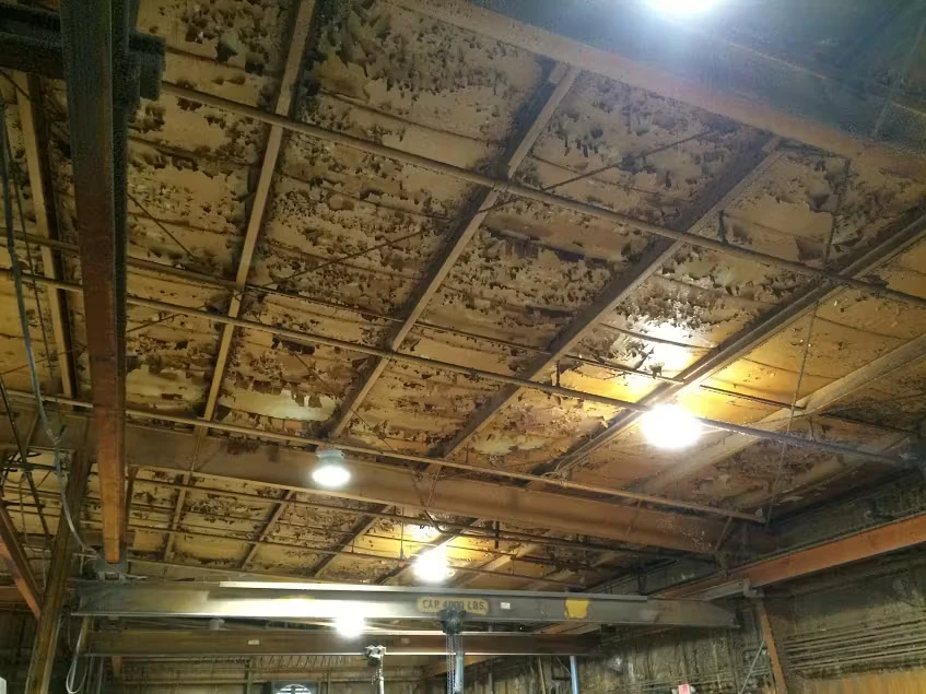 Interior of an industrial warehouse showing a grid of steel ceiling beams covered with peeling, flaking material; several bright hanging work lights and exposed wiring illuminate the space below.