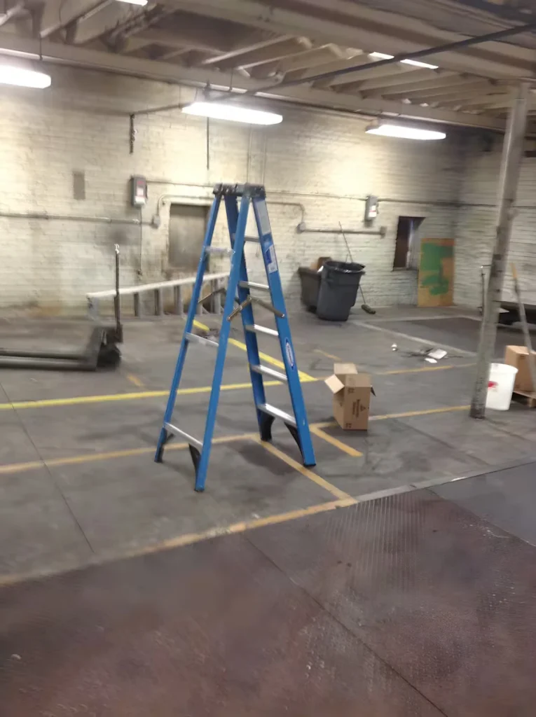 Industrial room with painted brick walls and fluorescent ceiling lights; a blue step ladder stands in the center near taped floor markings, with scattered boxes, a trash bin, and equipment along the edges of the concrete floor.