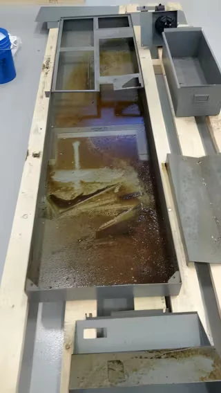 Long rectangular metal machine tray lying on pallets, partially filled with brown oily liquid and residue; removable metal panels and bins sit nearby on a clean workshop floor.