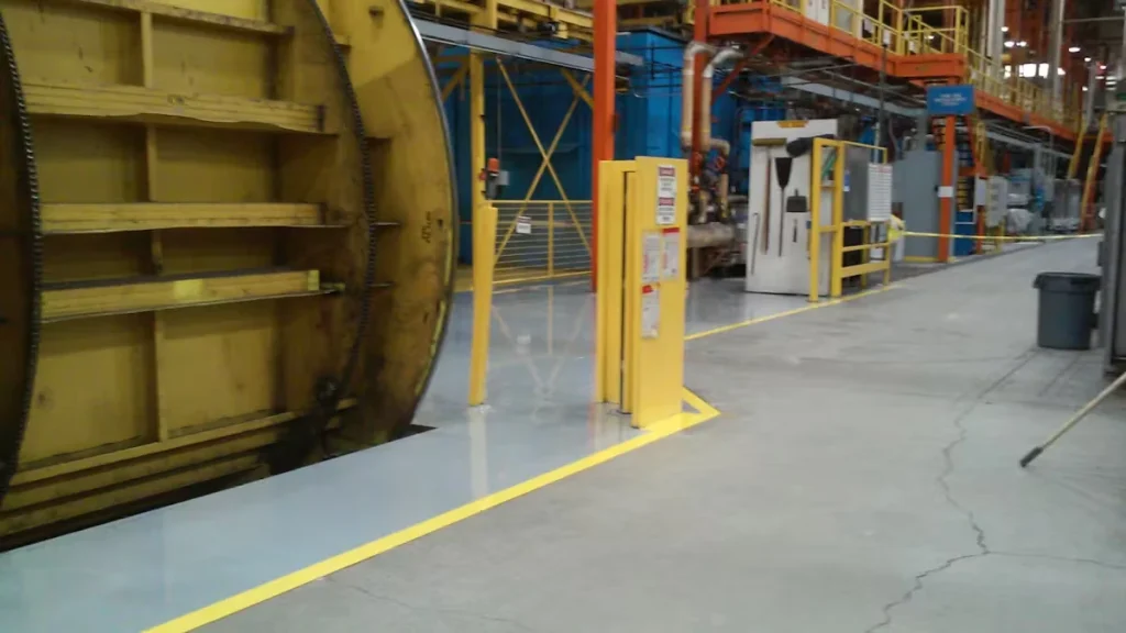 Factory floor with fresh gray epoxy coating and bright yellow safety lines marking walkways around large industrial machinery; guardrails, platforms, and equipment are visible in the background.