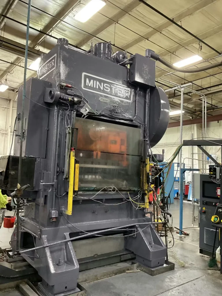 Large gray industrial press labeled “MINSTER” stands on a factory floor under fluorescent lights, with a protective front panel, exposed cables and hoses, and nearby control equipment in a manufacturing area.