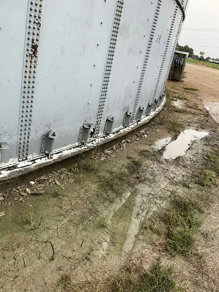 Close view of the curved base of a large riveted metal tank beside muddy ground with puddles; water stains and debris collect along the concrete foundation edge.