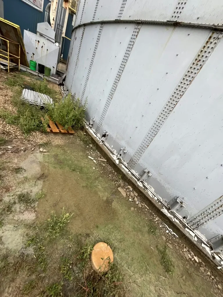 Lower section of a large riveted tank with darker staining near the base; exposed red-brown striping and multiple brackets line the curved foundation edge beside wet soil and small plants.