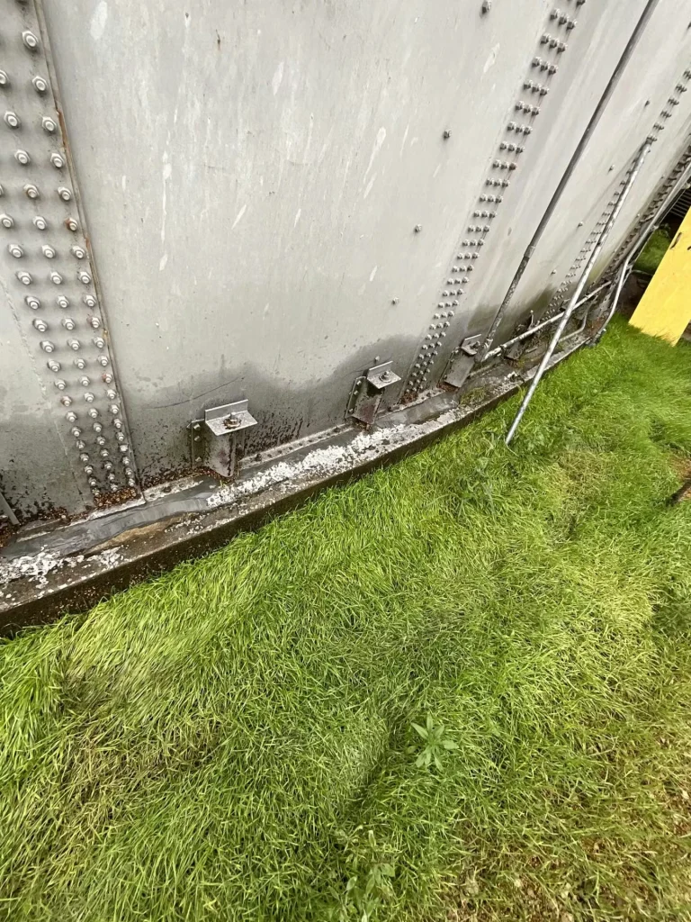 Lower wall of a large riveted metal tank above bright green grass; dark staining and chipped white coating run along the concrete base near several mounting brackets.