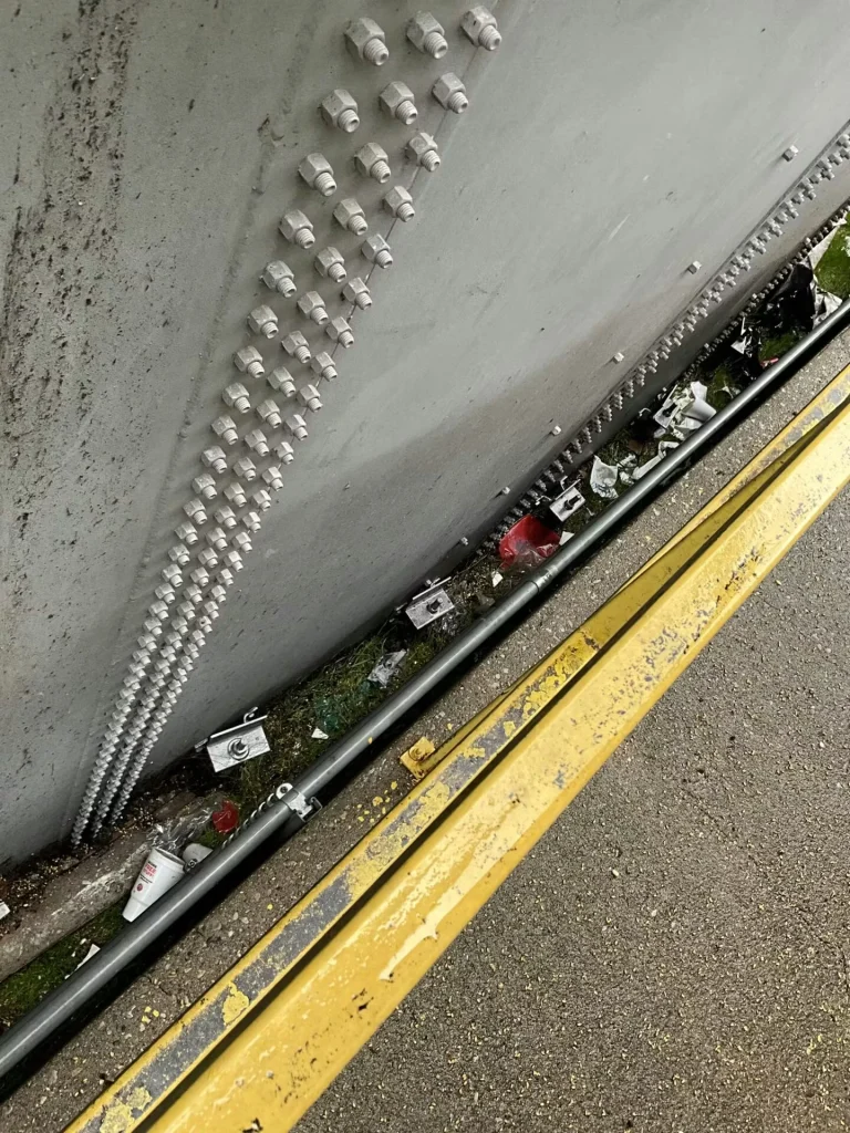 Tank base beside a gravel area with weeds and scattered debris; several rust-stained brackets are bolted to the tank wall above a worn sealant line on the concrete curb.