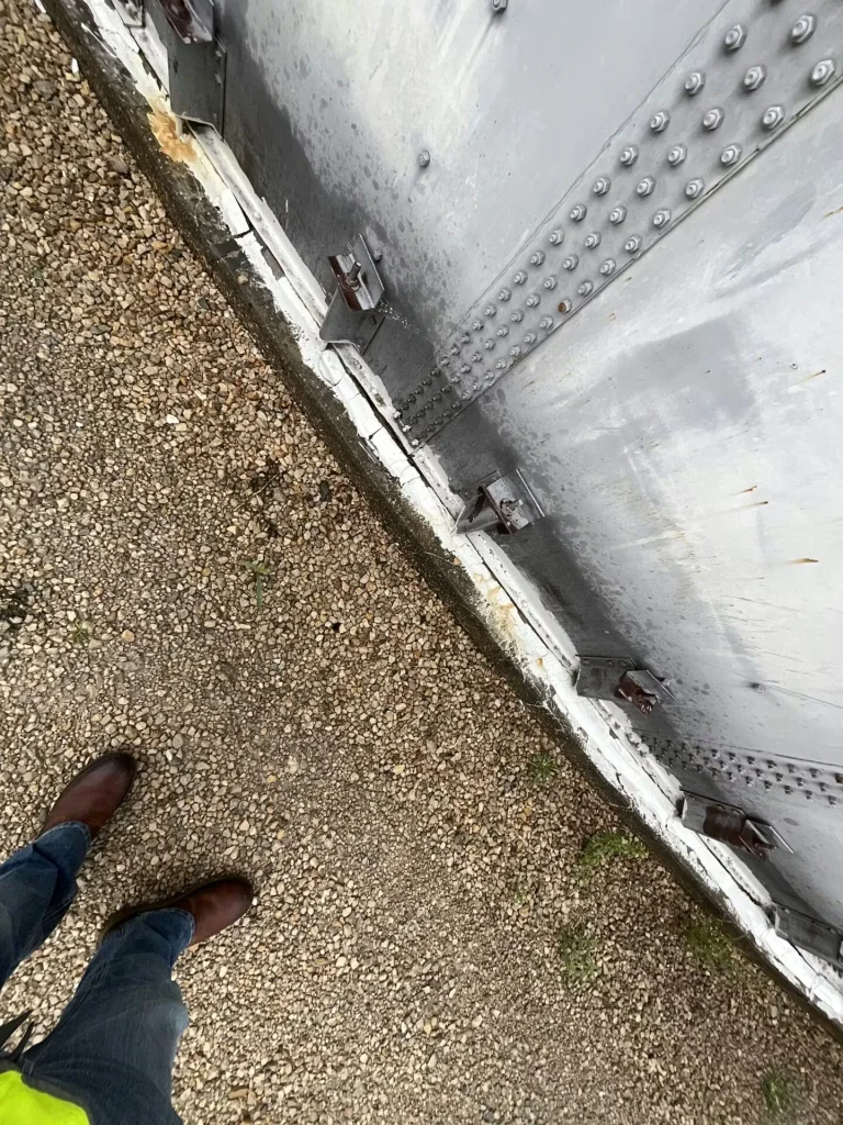 Tank wall with rows of rivets and multiple base brackets next to a patchy grassy area; an electrical box and conduit are mounted on the wall near nearby equipment.