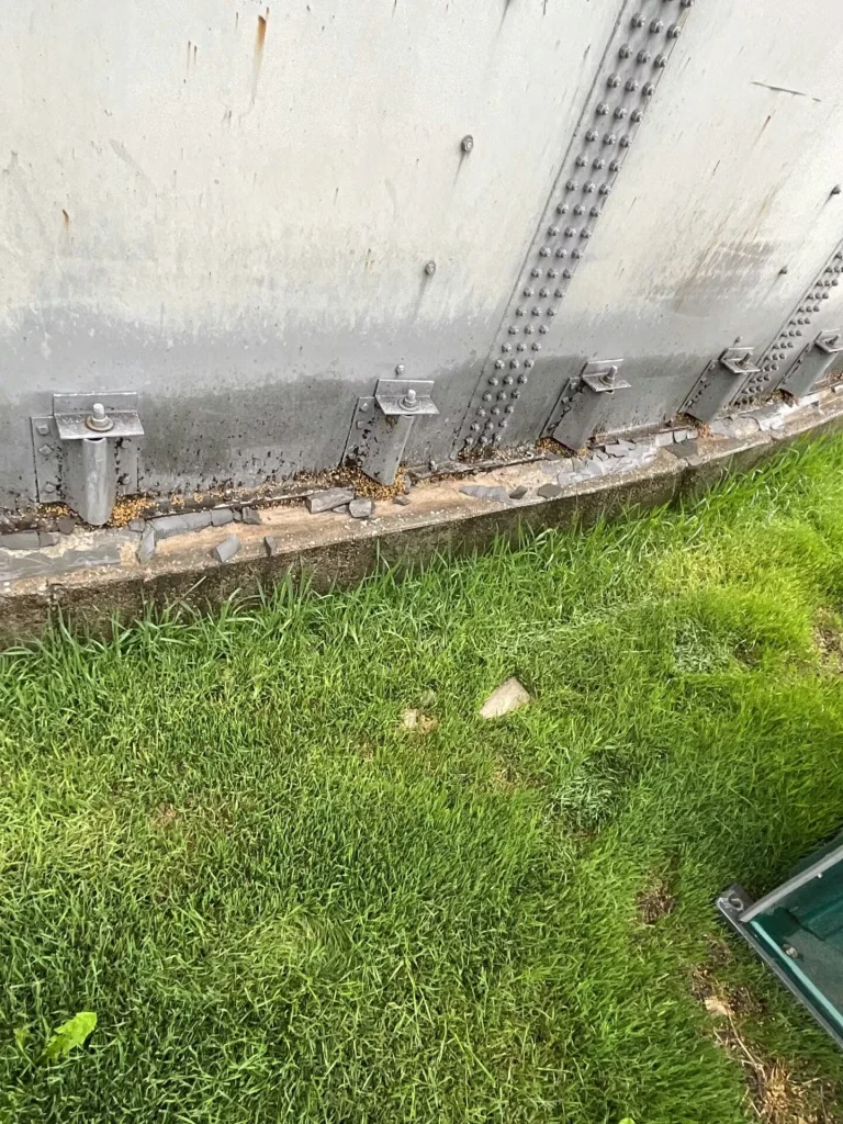 Close-up of a cracked and chipped concrete foundation edge at the base of a metal tank; loose stones and grime sit around a metal bracket and bolt.
