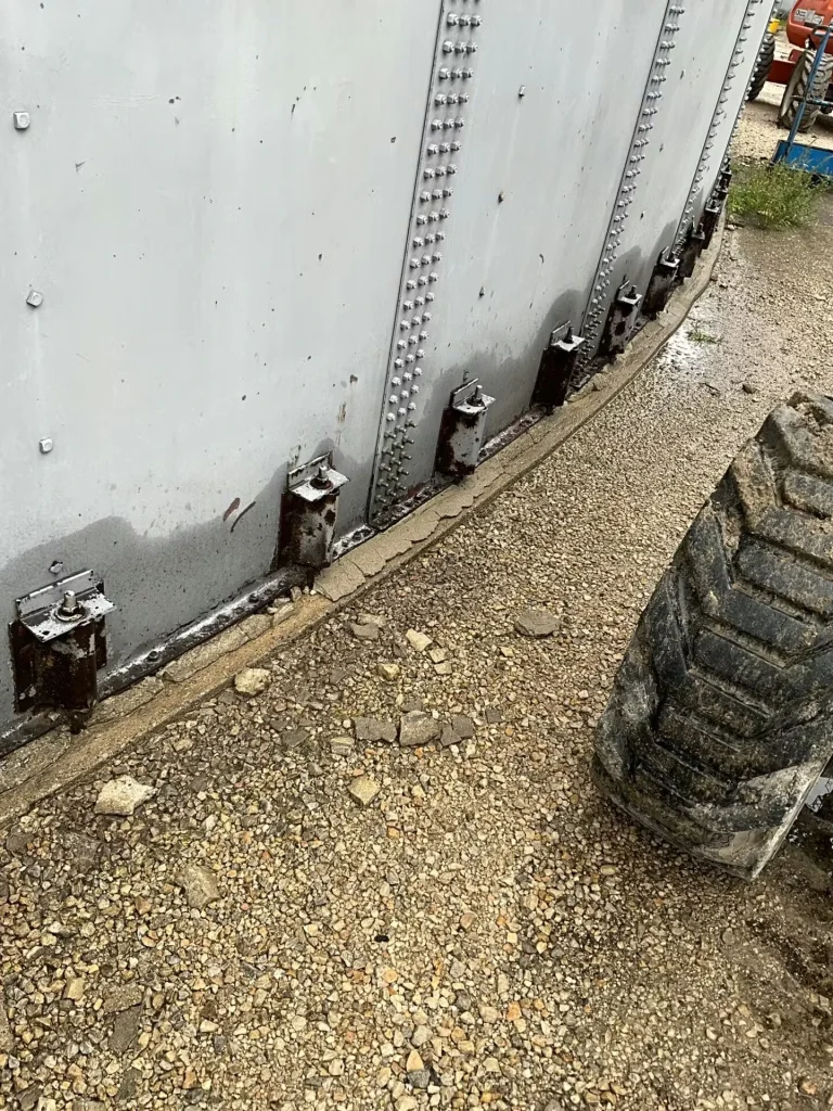Curved tank base along gravel, showing several dark, rust-streaked brackets and deteriorated sealant at the joint between the tank wall and concrete foundation; a large tire fills the right side of the frame.