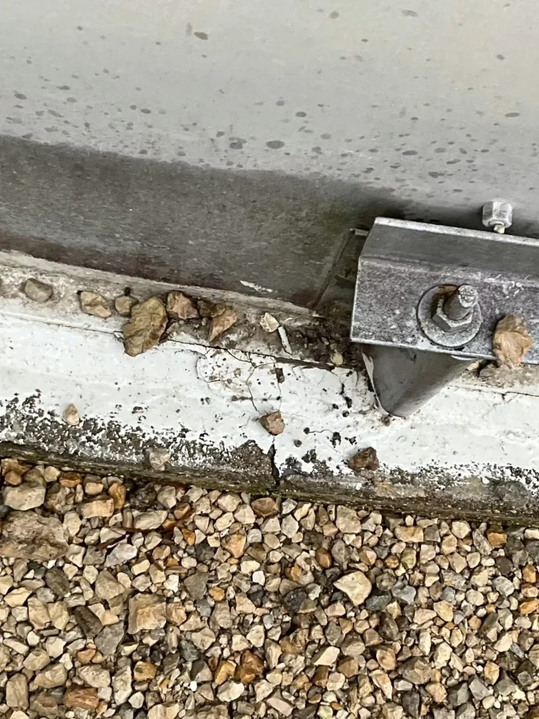 Tank base above grass with several brackets spaced along the concrete curb; the lower wall shows gray staining and scattered debris along the foundation edge.
