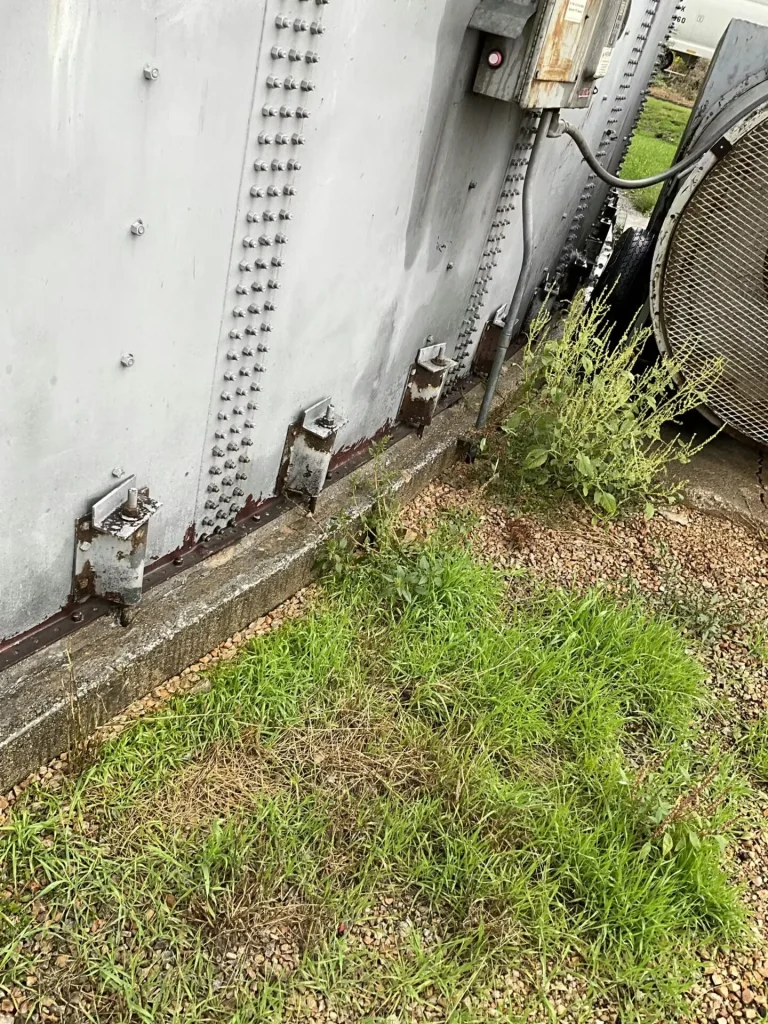 Angled view looking down at the tank base and gravel; a person’s boots and jeans appear at the left edge near a white-coated foundation seam and mounted brackets.