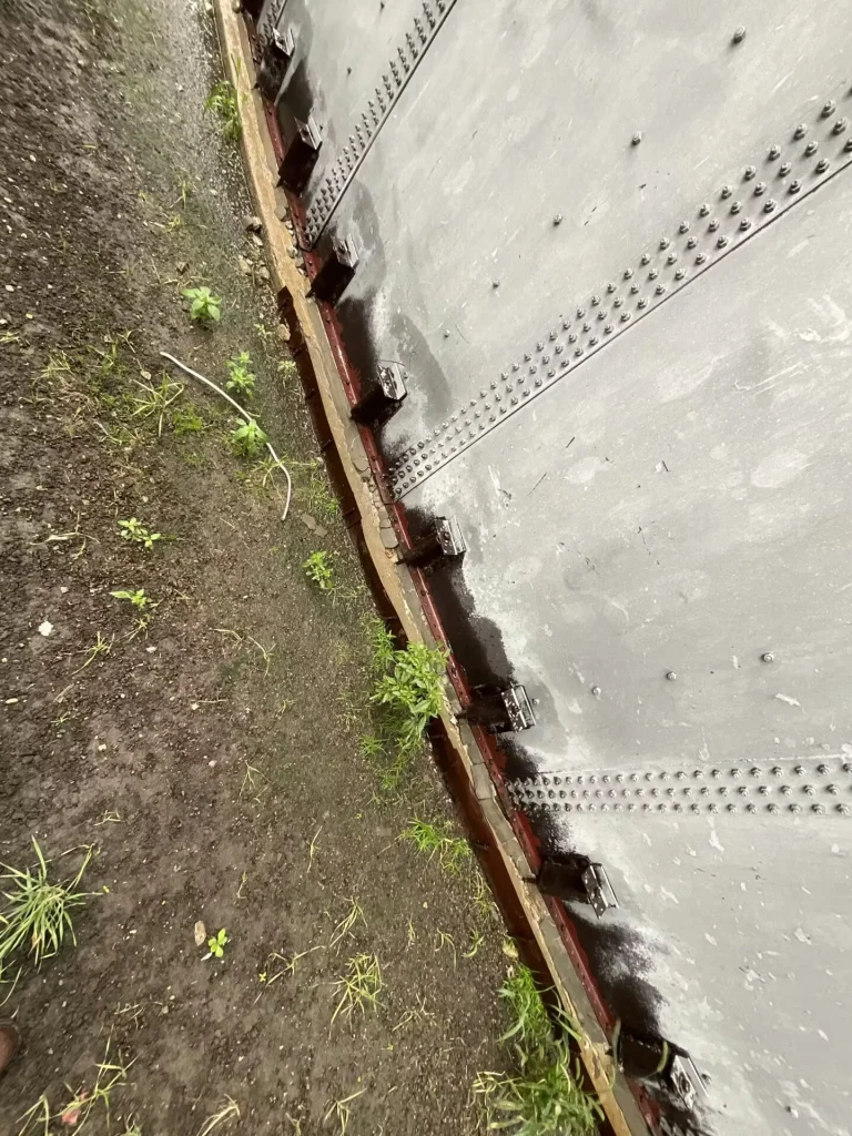 Angled view along the base of a large metal tank near gravel and weeds; standing water and algae-like discoloration appear around the foundation.