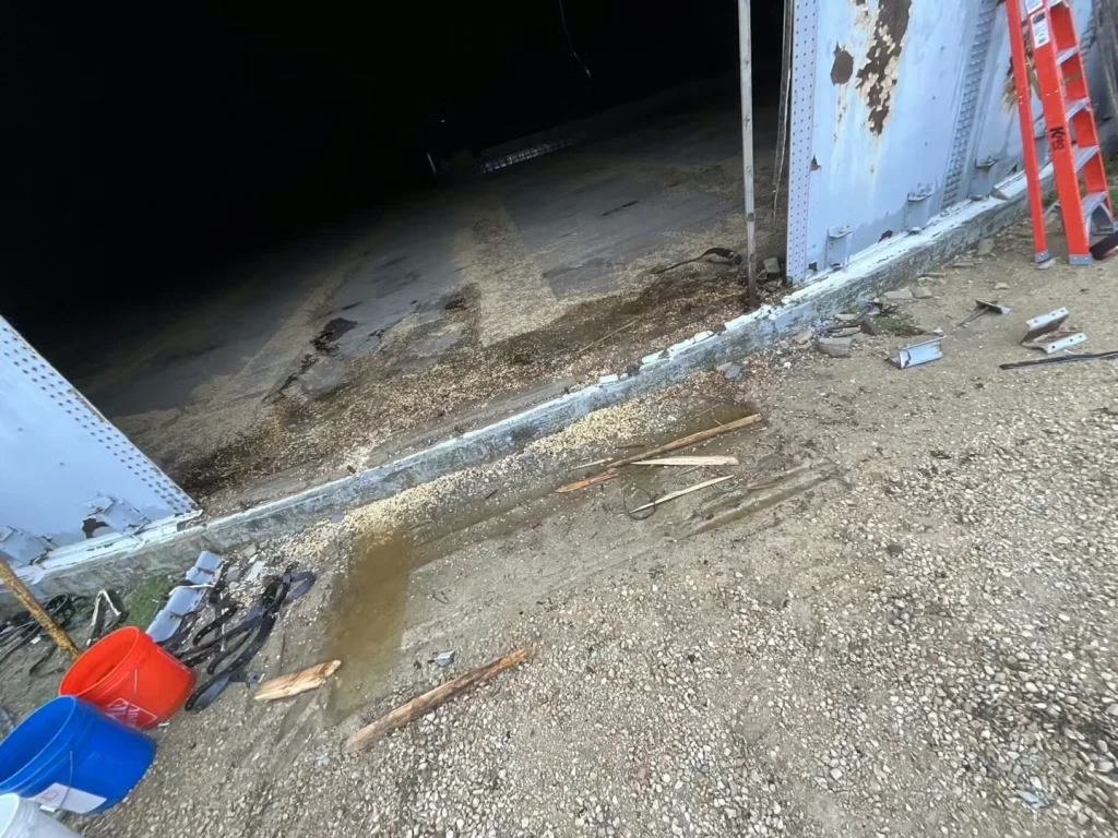 Close-up of tank wall rivets and mounting brackets above a concrete base; rust streaks and worn sealant are visible along the joint.