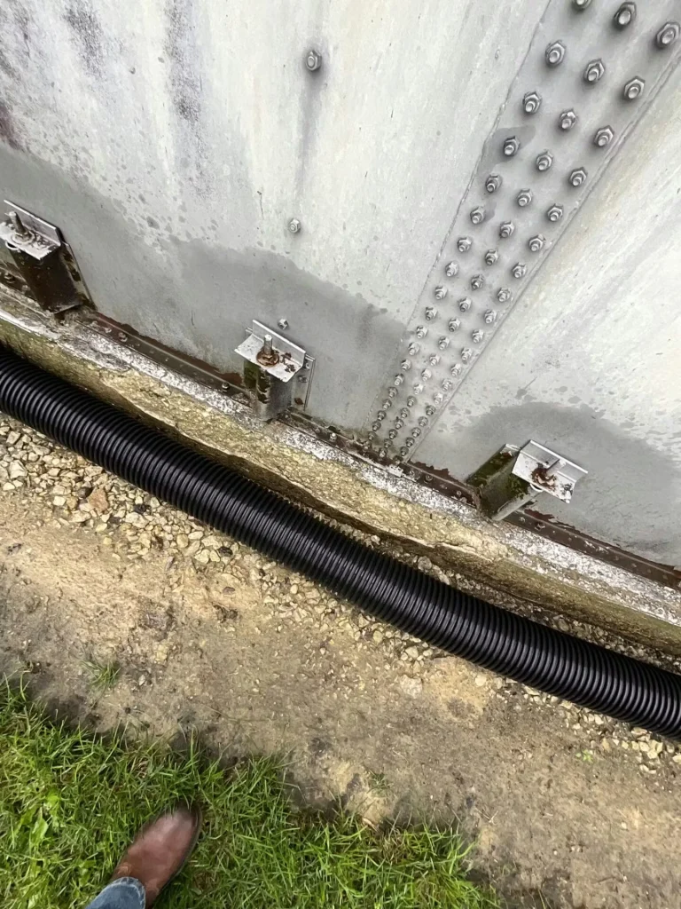 Detailed close-up of two metal brackets bolted to a tank wall above a concrete foundation; chipped coating and rust staining appear at the seam.