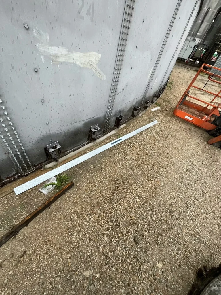 Tank base with a yellow metal panel leaning against the wall; loose debris, broken pieces, and a white rod lie on gravel near the concrete curb.