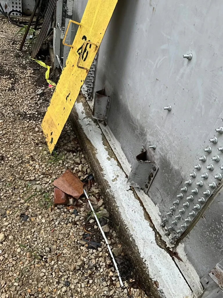 Wide view of the tank’s lower wall with multiple brackets along the base; a long straightedge lies on the gravel, and an orange lift platform sits to the right.