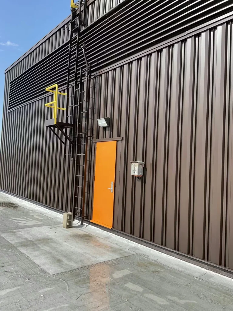 Rooftop penthouse wall with dark vertical metal siding; an orange door sits below a fixed ladder with yellow safety rails, under a clear blue sky.
