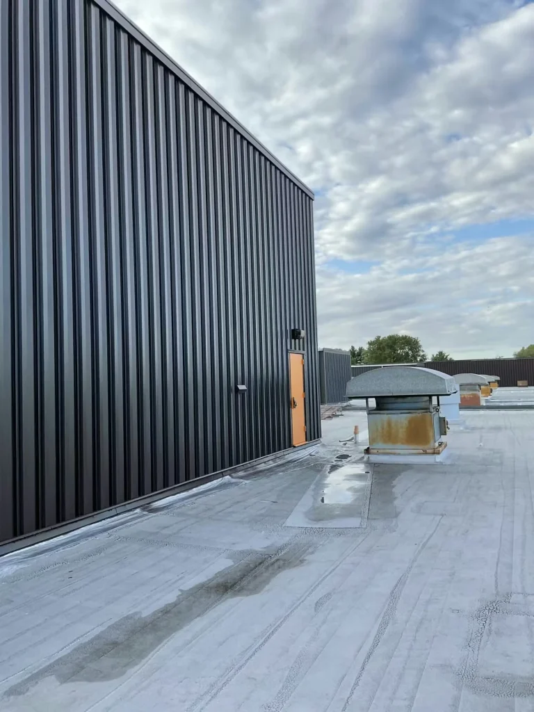 Rooftop view along the side of the penthouse; an orange door is visible in the distance, with roof equipment and a broad expanse of white/gray roof membrane.