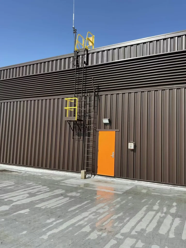 Front-on view of the rooftop penthouse with dark metal siding, orange door, and fixed ladder; buckets and tools are on the roof membrane in front.