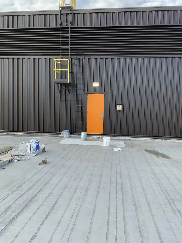 Rooftop penthouse wall with ladder and yellow safety rails; bright sun casts sharp shadows on the dark metal siding and roof membrane.