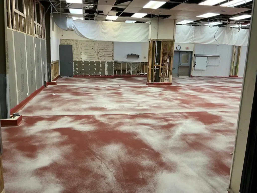 Wide view of a large indoor industrial space under renovation, with a red floor coating covered in scattered white powdery patches; exposed framing, plastic dust barriers, and ceiling tiles missing.