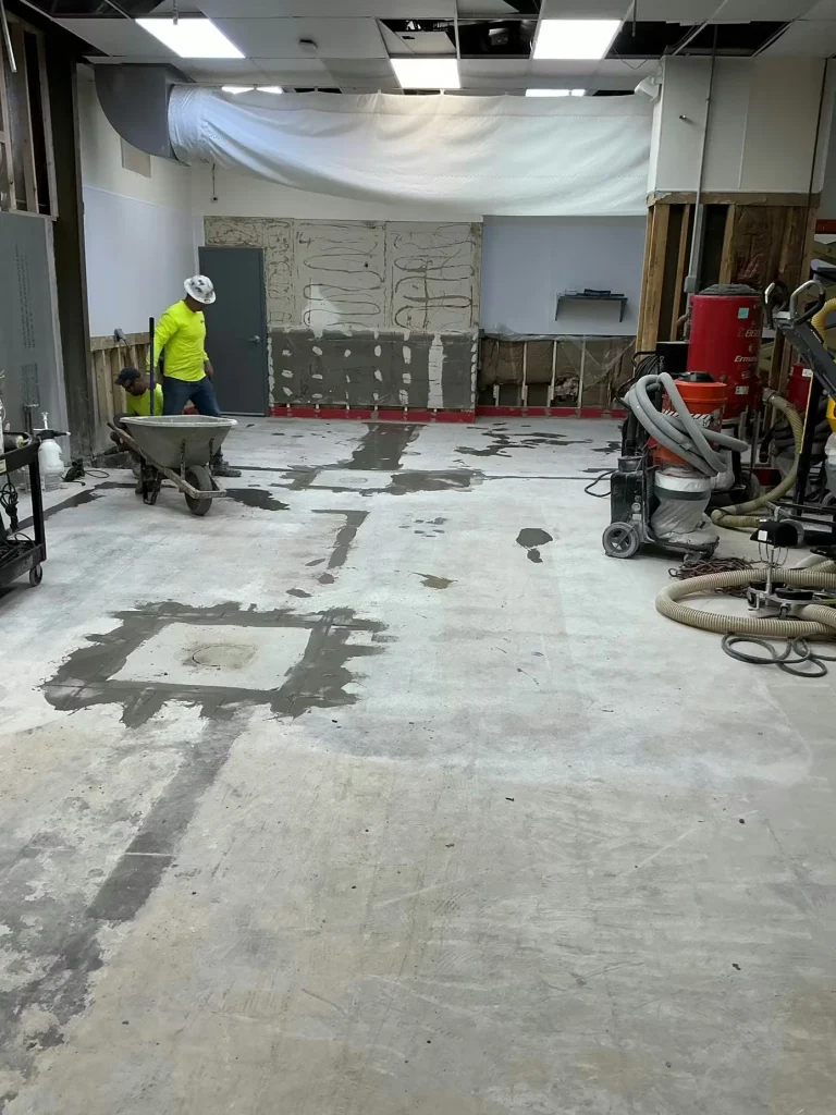 Large room with bare concrete floor showing dark repaired/filled areas; two workers in high-visibility shirts stand near a wheelbarrow, with construction tools and a vacuum system along the right side.