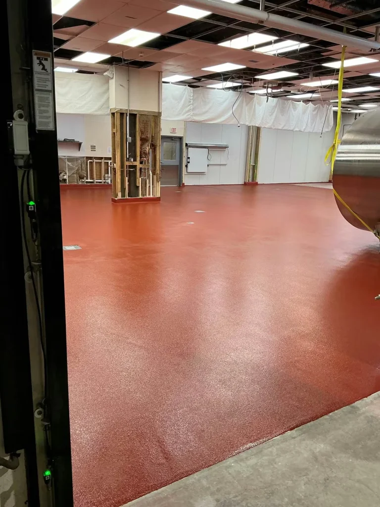 View from a doorway into the renovated space with a smooth, glossy red-coated floor; open ceiling grid with missing tiles and hanging cords, and a large metal tank partially visible on the right.