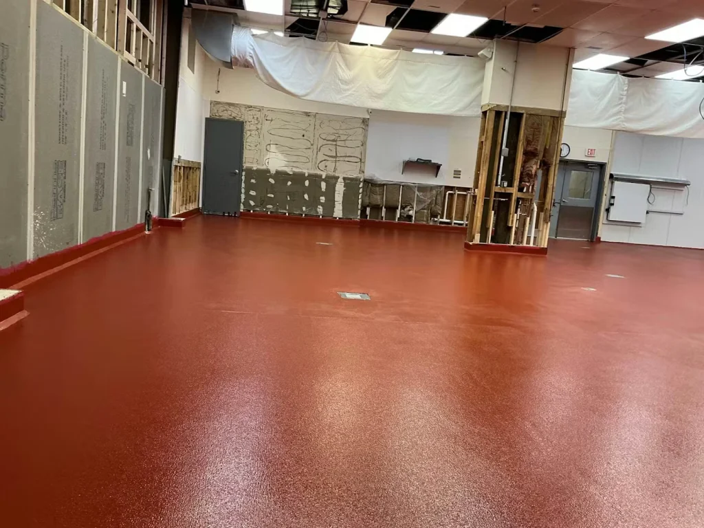 Same room after finishing, showing an even, glossy red floor coating extending across the space; exposed studs and temporary plastic sheeting remain along the walls and ceiling.