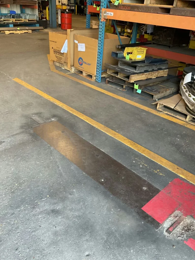 Warehouse floor with yellow lane lines and a dark rectangular patch in the concrete near the foreground; pallets, cardboard crates, and stacked metal plates sit under orange-and-blue industrial shelving.