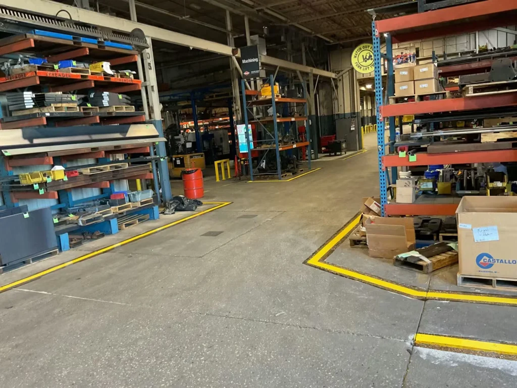 Factory floor with freshly painted bright yellow safety lines outlining walkways and a protected area around a column; a red STOP sign hangs above, with machinery and workstations visible in the background.