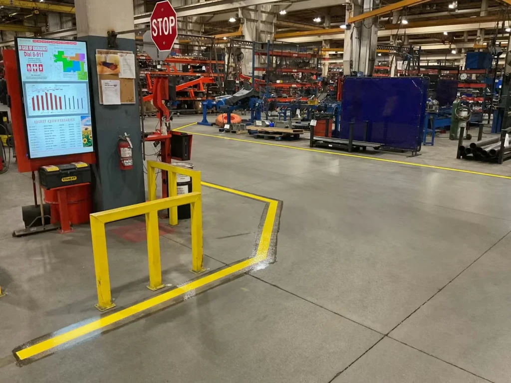 Wide view down a factory aisle bordered by crisp yellow floor striping; tall shelving on both sides holds metal stock and boxes, leading toward machining equipment deeper in the facility.