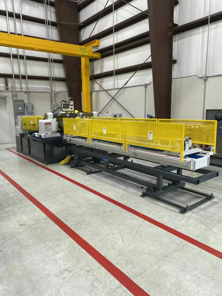 Long industrial machine on a shop floor, protected by yellow mesh safety guarding; red floor lines mark a boundary around the equipment, with a yellow overhead crane structure behind it.
