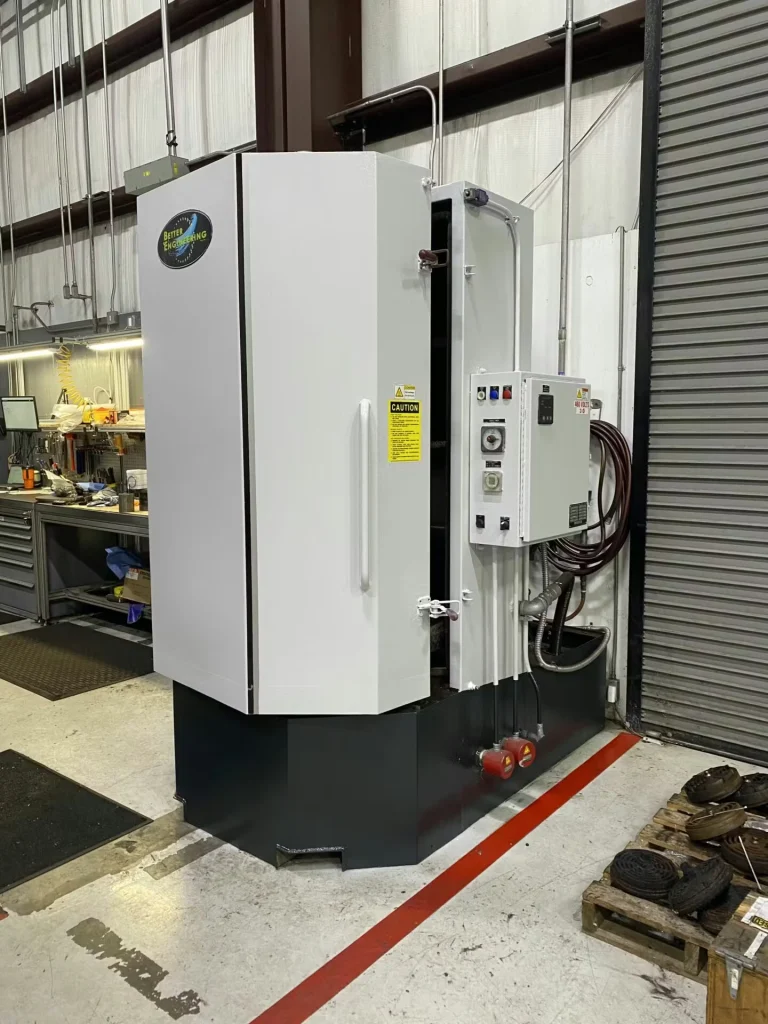 Manufacturing work area with two white workbenches holding circular metal parts and tools; red floor lines outline the workstation, with overhead piping, cables, and a mezzanine in the background.